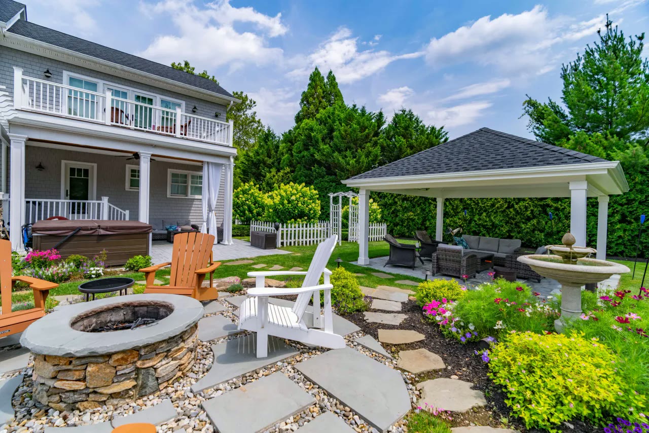 Stone fire pit and Adirondack chairs among flowering beds with a fountain and pavilion beyond