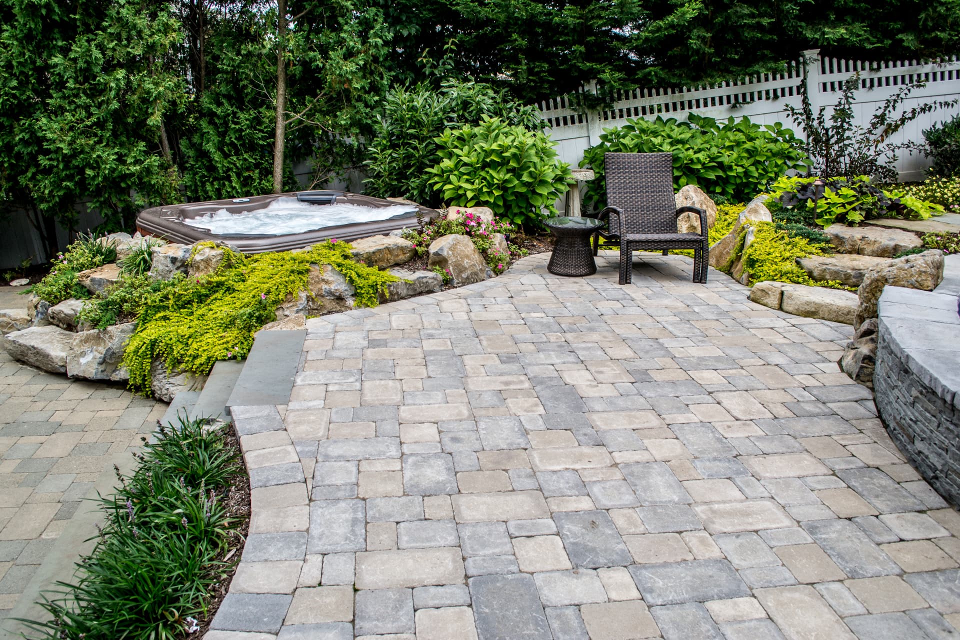 Paver patio leading to a hot tub set among boulders, planting and a lounge chair