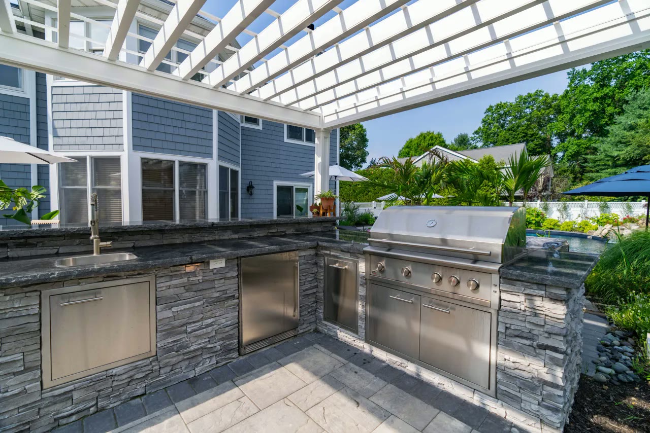 Stone outdoor kitchen with stainless grill, sink and refrigeration under a white pergola beside the pool