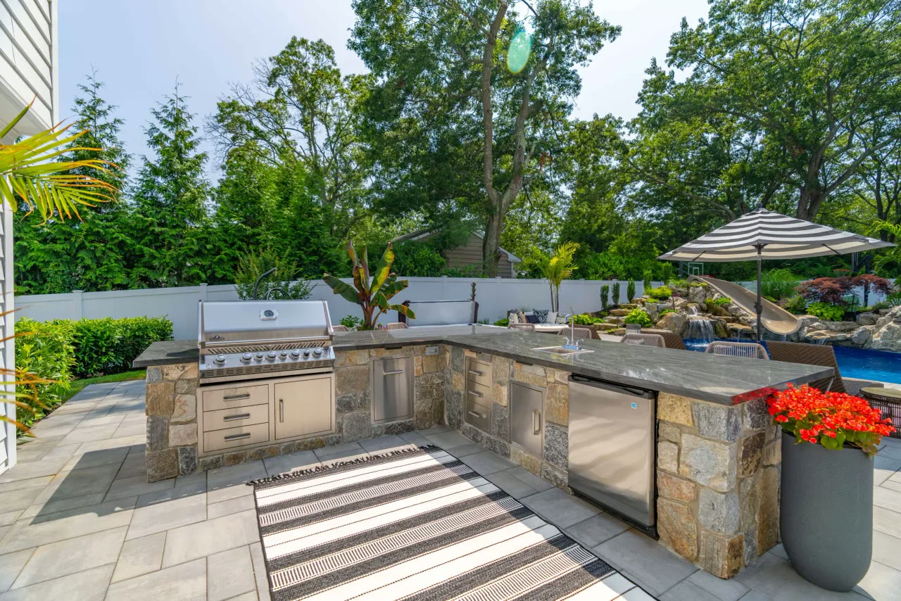 L-shaped stone outdoor kitchen with built-in grill, sink and bar seating overlooking a pool with waterfalls
