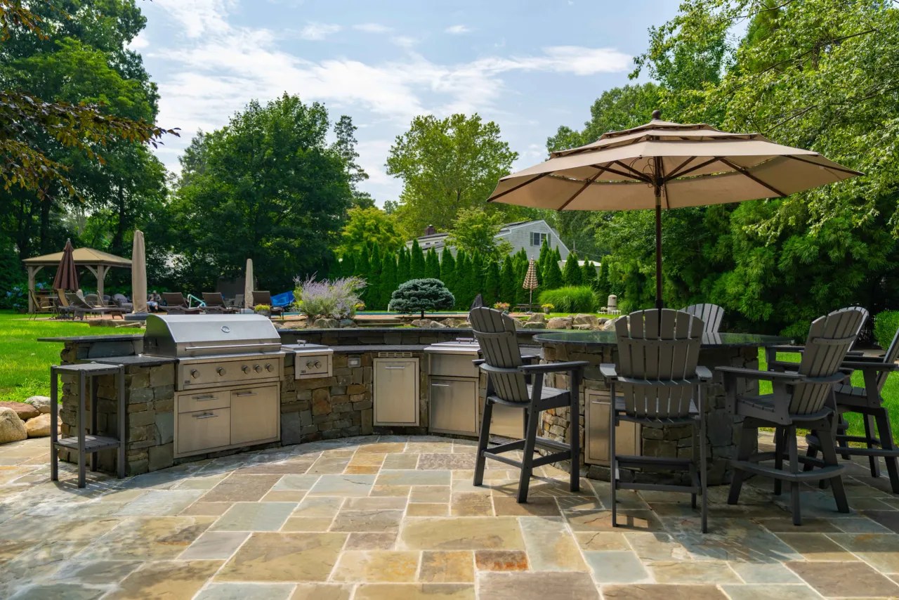 Outdoor kitchen with built-in stainless grill, side burner and bar seating under an umbrella