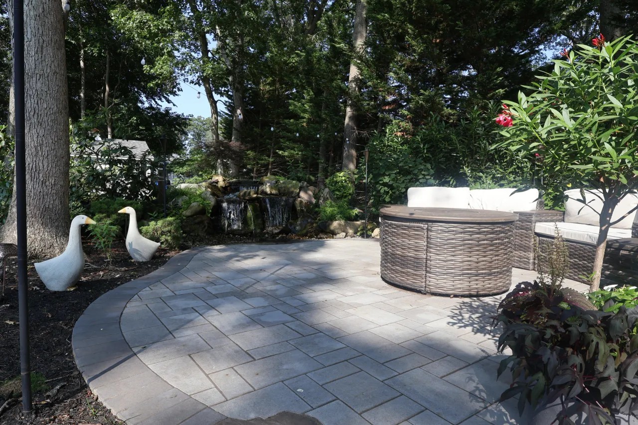 Curved gray paver patio with fire pit seating beside a pond waterfall