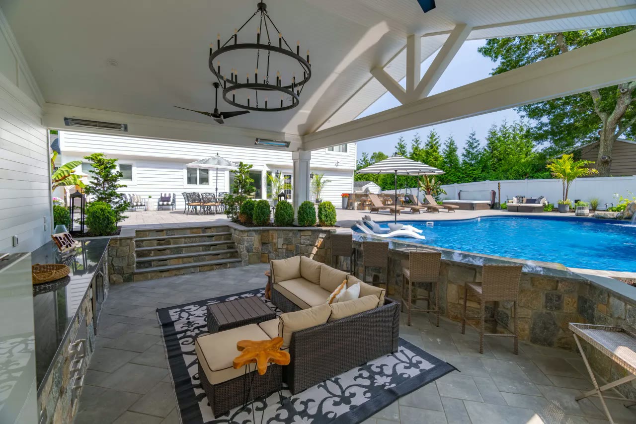 Covered outdoor lounge with chandelier, wicker seating and bar stools beside a pool