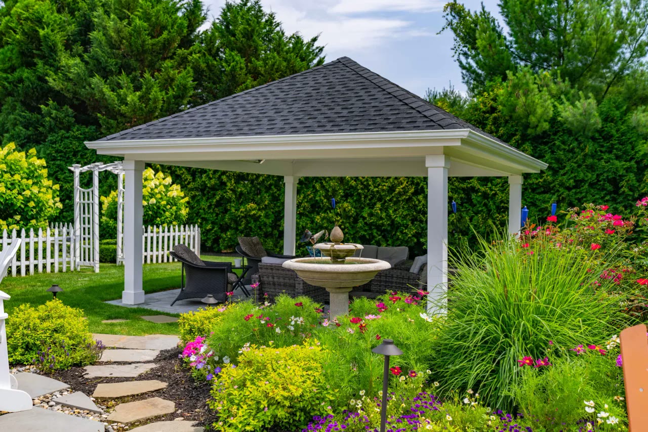 White pavilion with lounge seating and a garden fountain surrounded by flowering beds