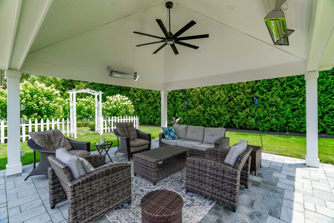 Inside a white pavilion with ceiling fan, heaters and wicker seating looking out to the garden