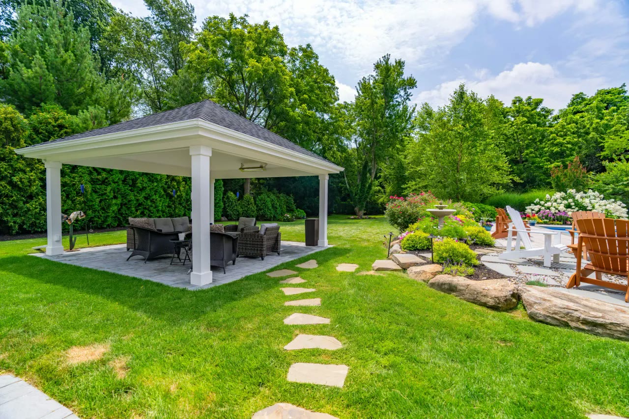 White pavilion with wicker seating beside a stepping-stone path and flowering garden beds