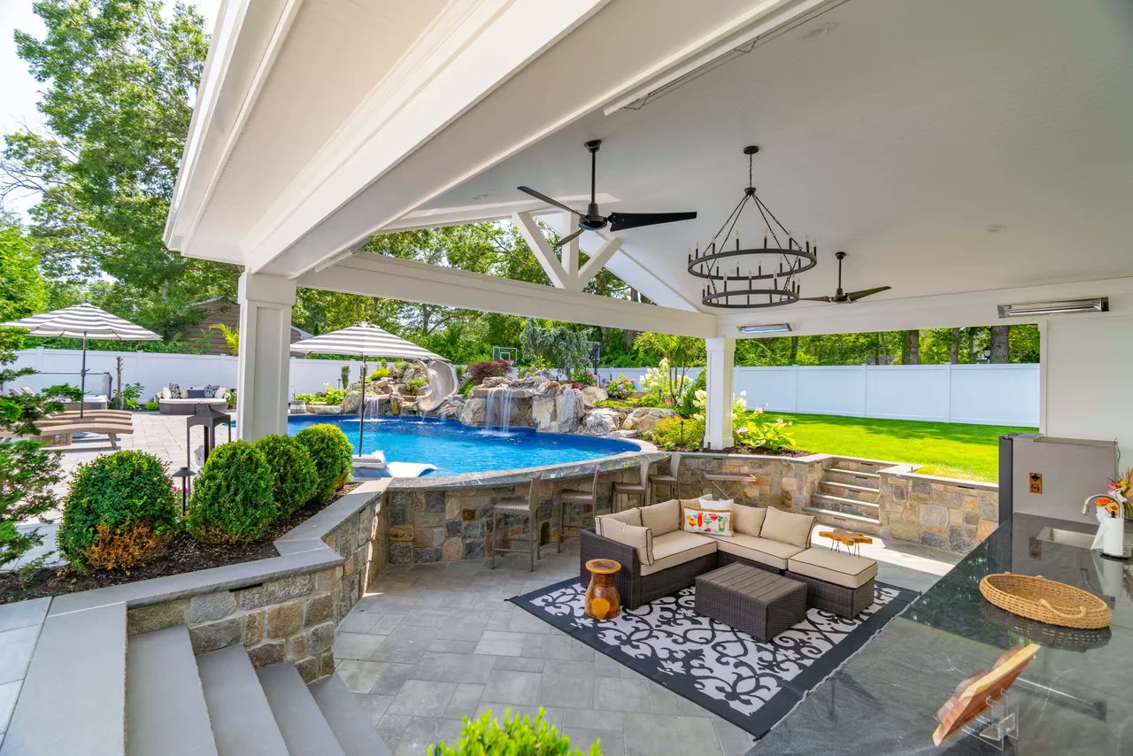 Open pavilion with lounge seating and outdoor kitchen facing a pool with rock waterfalls and a slide