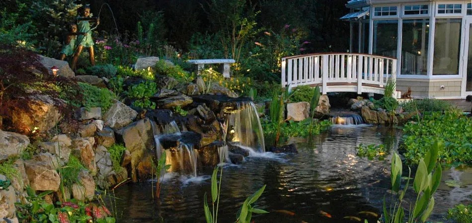 Illuminated pond waterfall and footbridge beside a glassed-in porch at dusk
