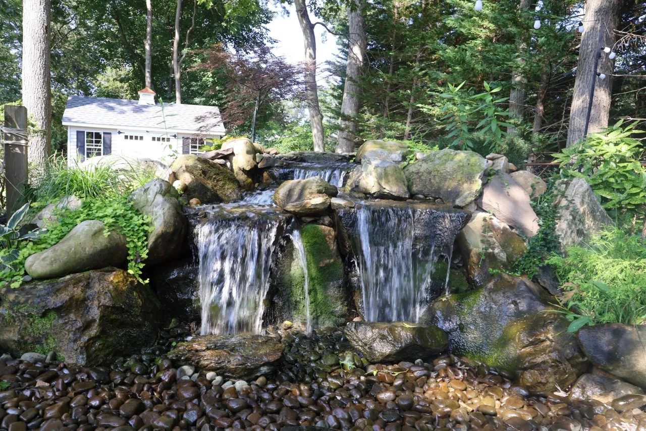 Twin mossy boulder waterfalls over a pebble beach beside a garden shed
