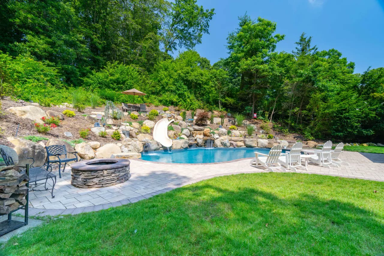 Freeform pool with slide and boulder waterfalls set into a planted hillside, fire pit and Adirondack chairs on the patio