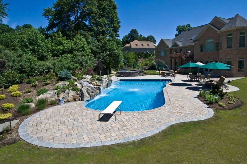 Freeform pool with diving board, deck jets and boulder waterfall inside a wide paver patio
