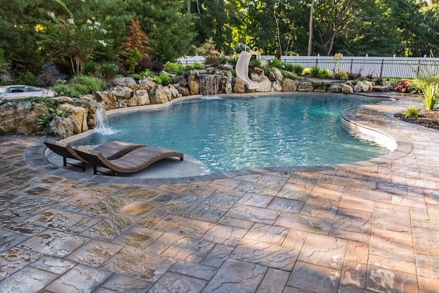 Freeform pool with slide and rock waterfalls beside wicker loungers on a paver patio