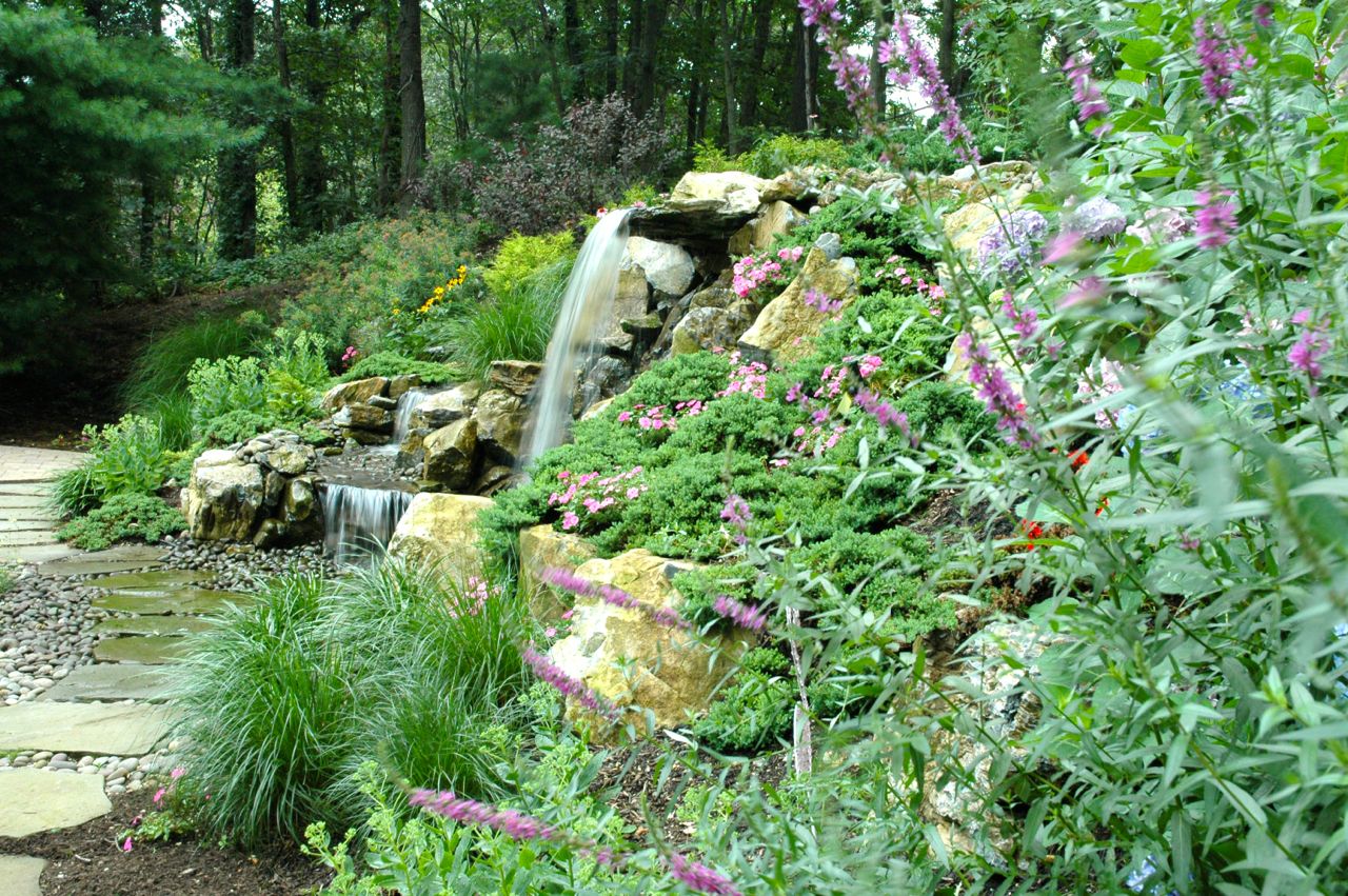 Natural Retaining Wall with Pondless Waterfall: Here, Deck and Patio used plantings as a key part of a natural retaining wall we built. “Along with boulders and other rocks, their root systems add to the overall strength of the support system,” says Dave Stockwell. “Deck and Patio always chooses plants for their color, bloom periods and how they grow. You can see the river rock we used here instead of concrete or asphalt so that the water seeps through into the reservoir below.